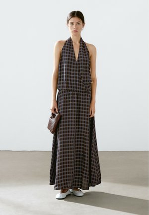 Robe maxi avec un col halter, présentant un motif à carreaux bleus et bruns. Tissu léger, taille cintrée et fentes sur les côtés. Ballerines blanches et sac en osier marron.