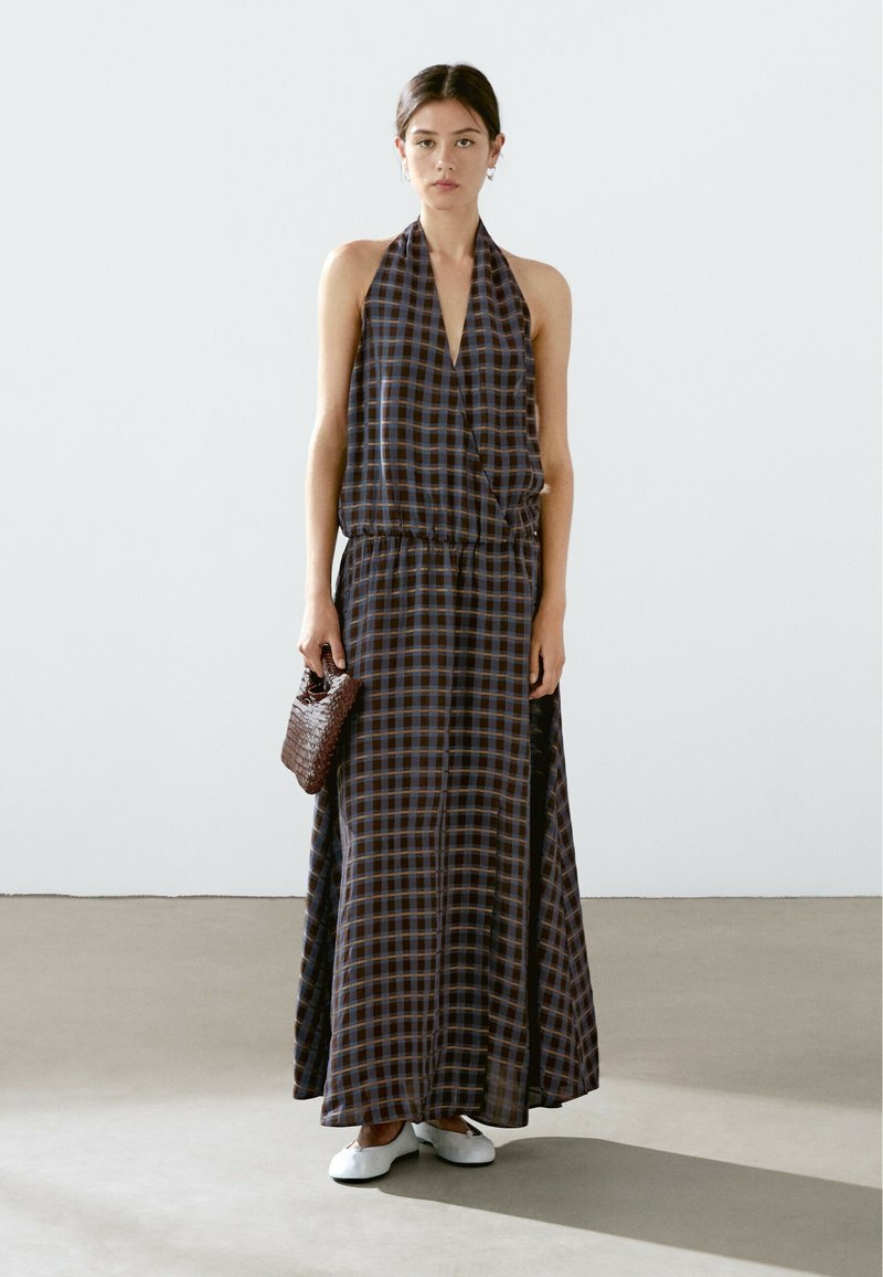 Maxi dress with a halter neckline, featuring a blue and brown checkered pattern. Lightweight fabric, cinched waist, and side slits. White flats and a woven brown bag.