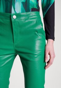 Gröna läderbyxor med en glansig finish, hög midja och en silverknapp accent framtill. Slät textur och åtsittande design.