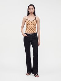 Jeune femme portant un caraco pêche avec des motifs de croissants noirs, un jean noir avec des coutures blanches et des sandales noires à talons, debout sur un fond blanc.