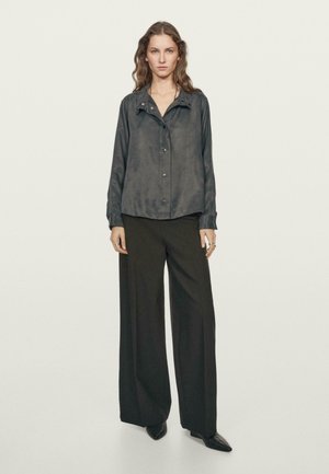 Camicia nera di seta con colletto e bottoni nascosti, abbinata a pantaloni neri a gamba larga e scarpe nere a punta, su uno sfondo chiaro.