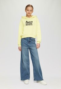 Gelber Kapuzenpullover mit "hey BENICE" Grafik, kombiniert mit weit geschnittenen blauen Streifenjeans und weißen Sneakern. Glattes Material, lässiges Design.