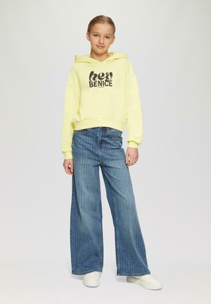 Żółta bluza z kapturem z grafiką "hey BENICE", w połączeniu z szerokimi niebieskimi spodniami w paski i białymi sneakersami. Gładki materiał, casualowy design.