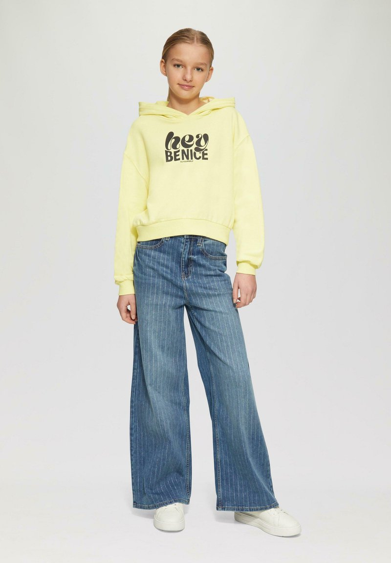 Gelber Kapuzenpullover mit "hey BENICE" Grafik, kombiniert mit weit geschnittenen blauen Streifenjeans und weißen Sneakern. Glattes Material, lässiges Design.