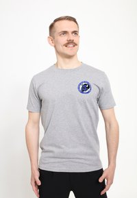 Grå bomulds t-shirt med korte ærmer og rund halsudskæring, prydet med en blå og sort grafisk logo på venstre brystområde.