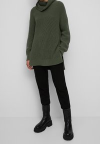 Personne portant un pull vert tricoté oversize, un pantalon noir et des bottes plateformes noires épaisses, debout devant un fond uni.