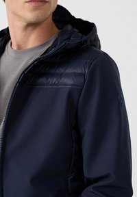 Marineblauwe hoodie met een gewatteerd bovenstuk, een gladde textuur, ritssluiting en logoprint op de mouw. Gdragen over een grijze shirt.