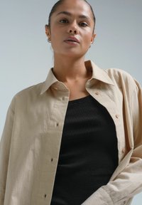 Hele beež button-up särk, mis on valmistatud pehmest ja tekstuurist kangast, on kihitatud sobiva musta ribakujulise topi peale. Nupud on kontrastse värviga.