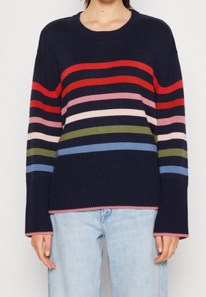 Granatowy sweter z dzianiny z poziomymi paskami w kolorach czerwonym, różowym, kremowym, oliwkowozielonym oraz jasnoniebieskim, noszony z jasnoniebieskimi dżinsami.