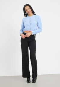 Ljusblå, kort cardigan med strukturerad stickning och knappar, kombinerad med svarta, rakbenade byxor och spetsiga svarta klackar mot en neutral bakgrund.