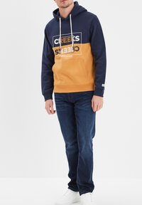 Sudadera con capucha de diseño en bloques de color azul marino y mostaza, que incluye un bolsillo canguro frontal y texto gráfico. Cuerdas blancas acentúan la capucha.