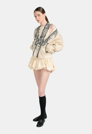 Donna che indossa un mini abito beige gonfio e giacca abbinata con ricamo di drago nero, abbinati a calzini neri al ginocchio e mocassini neri.
