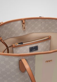 Beige tote bag met tan leren accenten, uitgerust met een gepatterneerde binnenzijde, een ritsvak en een logo-label. De binnenkant is glad en ruim.