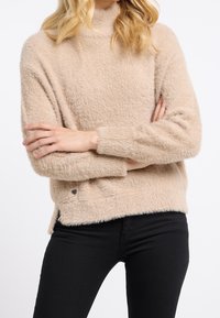 Beige, fluffig tröja med hög krage. Har en strukturerad yta, långa ärmar och en sidoklyvning vid fållens nederkant. Kombineras med svarta byxor.