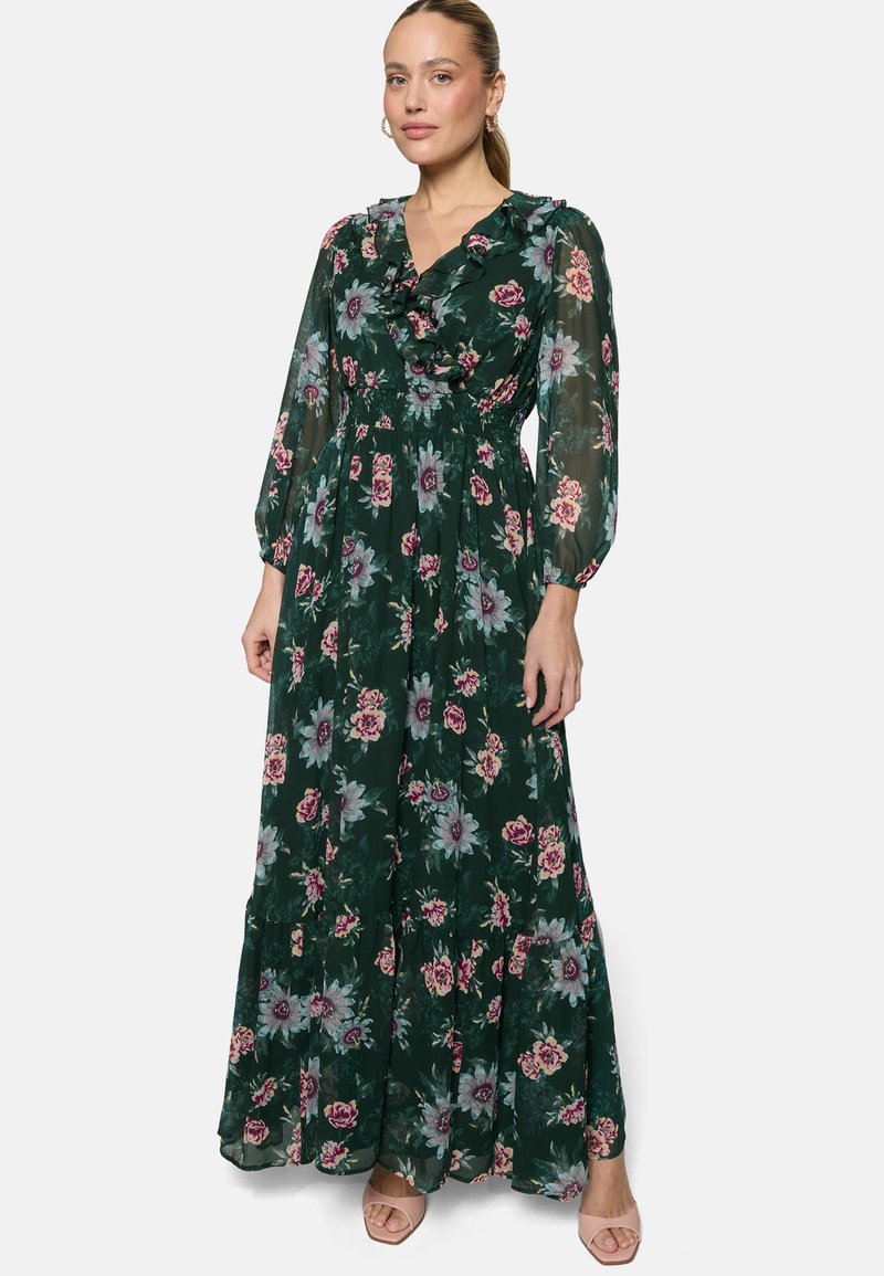 Femme portant une longue robe verte foncée à fleurs avec un col à volants et des manches longues transparentes, debout devant un fond uni.