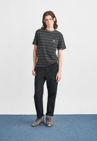 Černé a šedé pruhované tričko s krátkým rukávem a kapsou na prsou, spárované s černými kalhotami a šedými turistickými botami na modré podlaze.