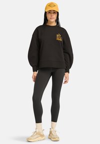 Schwarzer Pullover mit Puffärmeln und gelbem Timberland-Logo, kombiniert mit dunklen Leggings und cremefarbenen Sneakers mit strukturierten Akzenten. Gelbe Mütze auf dem Kopf.