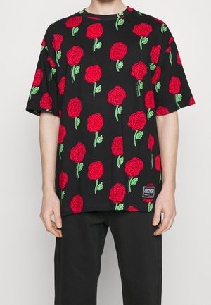 Person trägt ein schwarzes Kurzarmshirt mit einem roten Rosenmuster, grünen Blättern und einem kleinen Markenlabel nahe dem Saum.