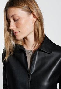 Giacca in pelle nera con colletto, apertura a zip e texture lucida. Indossata sopra un top grigio e testurizzato, che mette in mostra un design elegante.