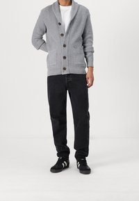Hombre vestido con un cárdigan gris abotonado, camiseta blanca, jeans negros y zapatillas negras con rayas blancas, de pie contra un fondo blanco liso.