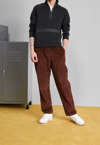 Sudadera de forro polar negra con media cremallera y bolsillo frontal, combinada con pantalones de pana marrones. Zapatillas blancas completan el conjunto.