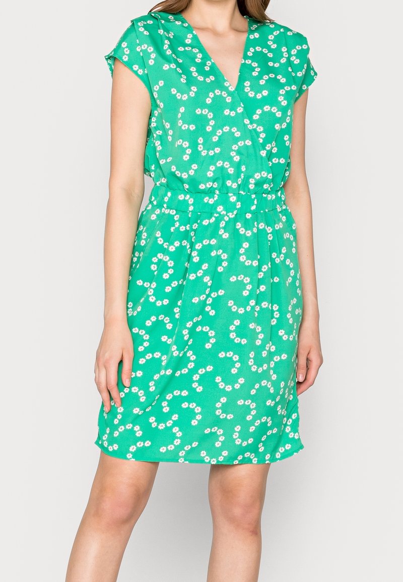Femme portant une robe verte à manches courtes avec un motif floral blanc et un col en V, debout devant un fond clair uni.