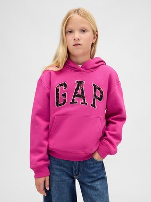 Blond kind dat een felroze hoodie met de letters "GAP" en een blauwe spijkerbroek draagt, staand met één hand in de zak tegen een effen achtergrond.