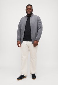 Chaqueta bomber gris claro con bolsillo con cremallera, camiseta negra, pantalones crema y zapatillas negras con suelas de goma. Texturas suaves en todo.