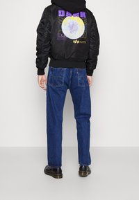 Czarna kurtka bomber z grafiką na plecach, zestawiona z niebieskimi jeansami o prostych nogawkach. Buty są czarne z grubą podeszwą.
