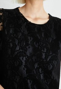 Top in pizzo nero con motivi floreali, scollatura rotonda e maniche corte. La trama è trasparente con una morbida fodera sottostante.