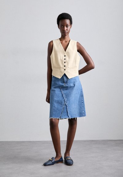 Beige linnen vest met knopen, gecombineerd met een blauwe denimrok met een voorzicht en een rafelrand, gedragen met marineblauwe instappers.