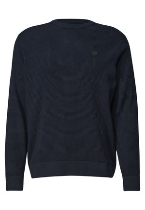 Homme aux cheveux foncés et à la barbe, portant un pull bleu marine et un jean bleu clair, assis contre un mur blanc avec un genou relevé.