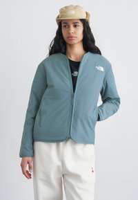 Νεαρή γυναίκα που φοράει τζάκετ με φερμουάρ σε τιρκουάζ απόχρωση, μπεζ καπέλο με επένδυση για τα αυτιά, λευκό παντελόνι και μαύρο πουκάμισο με ορατά λογότυπα στο τζάκετ και στο καπέλο.