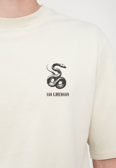 T-shirt en coton crème arborant un design de serpent noir avec le texte "HAN KJØBENHAVN" en dessous. Manches courtes et encolure arrondie.