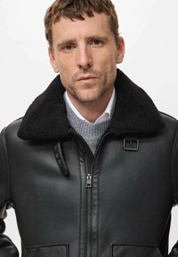 Chaqueta de cuero negra con un cuello de borreguito negro, cierre frontal con cremallera y bolsillos laterales, llevada sobre un suéter gris y una camisa blanca.