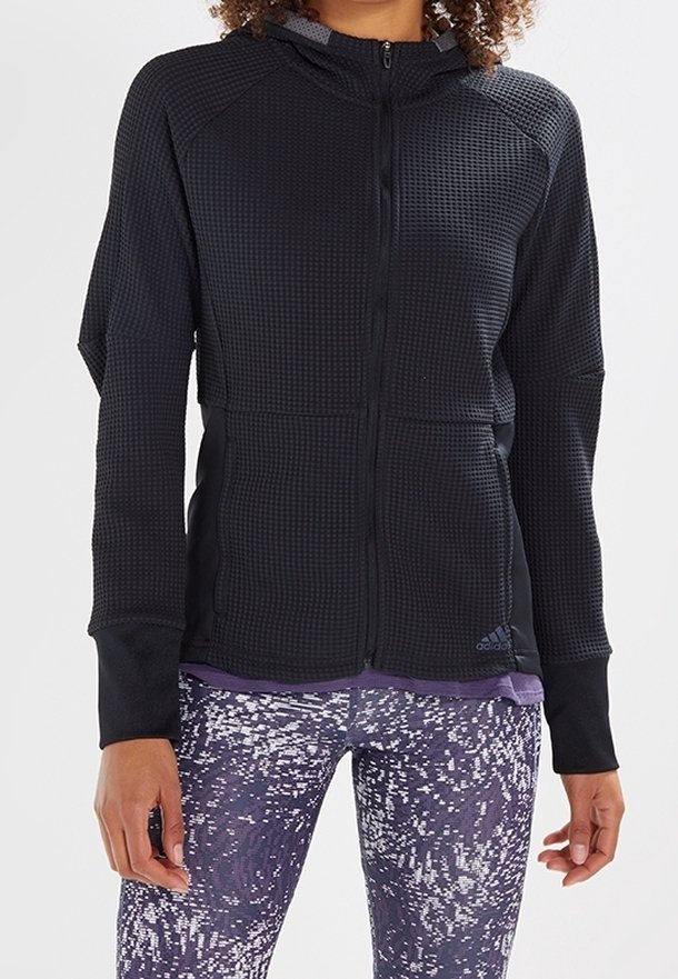 Veste d'entraînement noire à fermeture éclair avec tissu texturé et capuche. Elle possède des manches raglan, des orifices pour les pouces et le logo Adidas. Associée à des leggings à motifs.