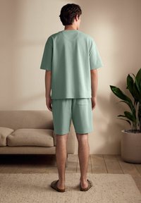 Lys grøn, t-shirt i bomuldsblanding med korte ærmer og matchende shorts. Afslappet pasform, enkelt design. Person står på et tekstureret tæppe.