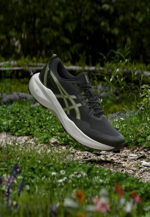 Zapatilla de correr negra y verde oliva con suela blanca, flotando sobre un camino rocoso de jardín con plantas verdes y flores alrededor.