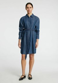 Sininen farkkukleid, jossa on napitettavat kaulukset, pitkät hihat ja kangasvyö vyötäröllä. Sileä pinta. Matteat mustat kengät.