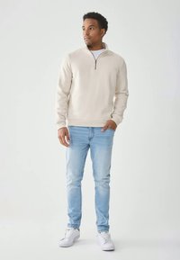 Férfi áll, bézs színű negyed-zip pulóverben, világoskék farmernadrágban és fehér tornacipőben, balra néz, az egyik kezét a zsebében tartva.