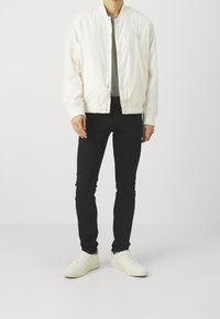 Chaqueta bomber blanca con botones a presión, camiseta gris debajo, jeans slim-fit negros y zapatillas blancas. Diseño simple y textura suave.