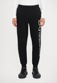 Svart joggebukse med elastisk midje og smalnende ben. Har hvit trykt logo langs venstre side. Laget av mykt bomullsblandingsmateriale.