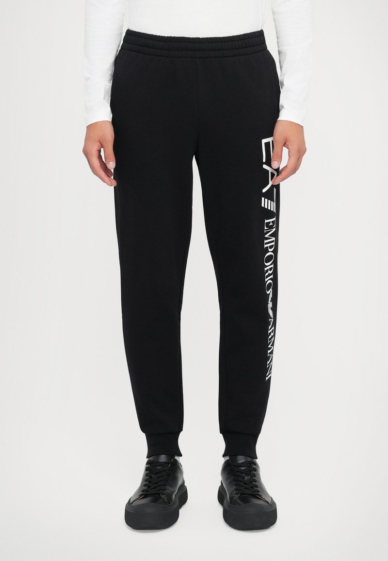 Svart joggebukse med elastisk midje og smalnende ben. Har hvit trykt logo langs venstre side. Laget av mykt bomullsblandingsmateriale.