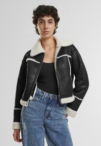 Veste en simili cuir noir avec col en fausse fourrure blanche et détails, design court, fermeture éclair, portée sur un haut noir et un jean bleu clair.