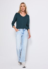 Donkerblauwe lange mouwen top met een ronde hals, gecombineerd met lichtblauwe wijde jeans. Draagt witte en grijze sneakers, poserende tegen een effen achtergrond.