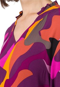 Blouse à manches longues avec un col en V, présentant un motif abstrait coloré dans des tons de violet, rose, orange et gris. Détail de col froncé.