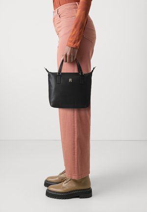 Person mit rosa Hose und braunen Schuhen, die eine schwarze Handtasche mit goldenem Logo vor einem schlichten weißen Hintergrund hält.