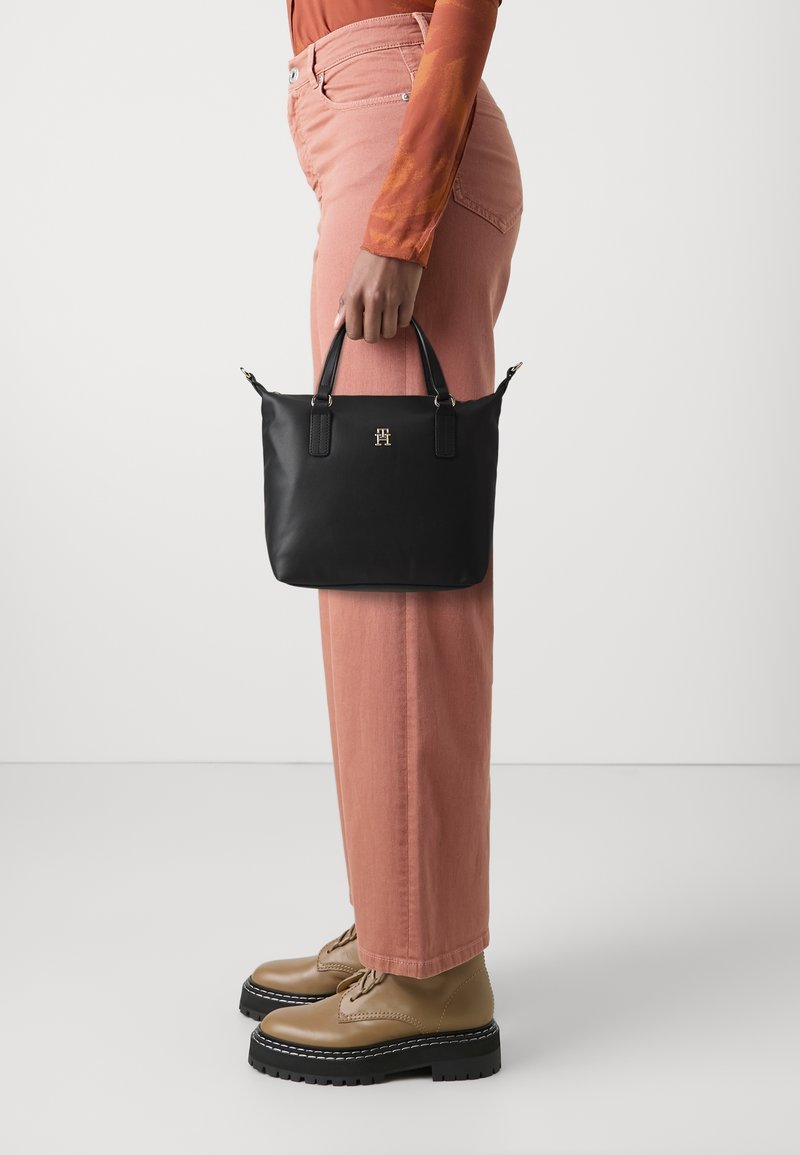 Osoba w różowych spodniach i brązowych butach trzyma czarną torebkę z złotym logo na jednolitym białym tle.