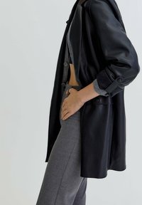 Manteau en cuir noir au design structuré, avec de grands boutons, un col châle et des manches retroussées, associé à un pantalon gris.