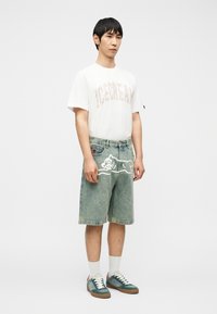 Νέος άνδρας που φοράει λευκό μπλουζάκι με τη λέξη "ICECREAM", τζιν σορτς με λευκό περίγραμμα σχεδίου ζώου, λευκές κάλτσες και μπλε-πράσινα αθλητικά παπούτσια, στέκεται ακίνητος.